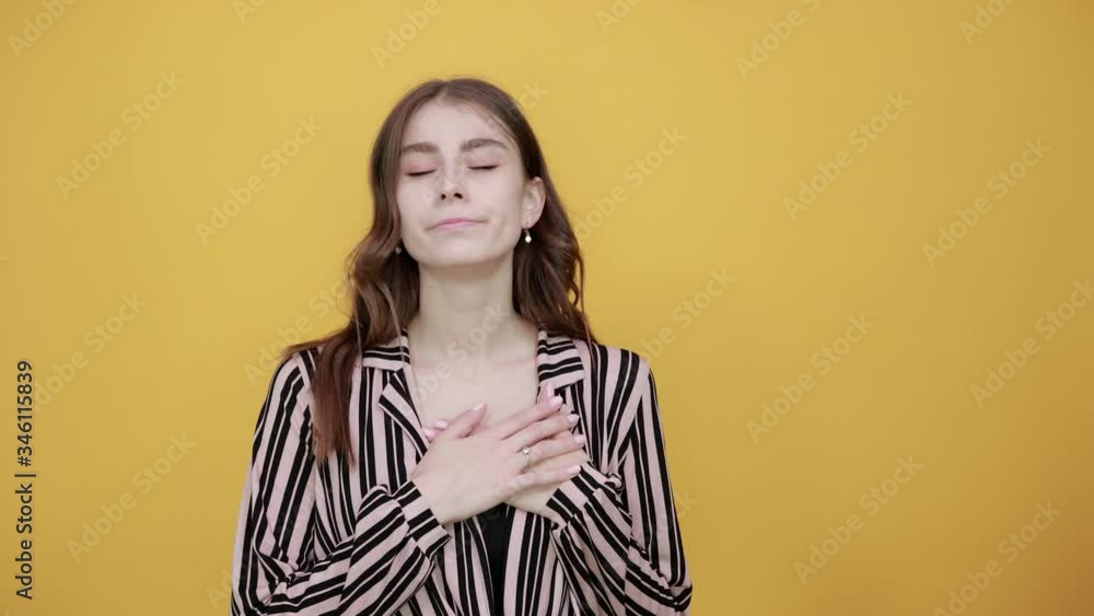Cute Young Woman Light Brown In Striped Pink, Black Shirt On A Yellow Background, Dreamy Girl Put Her Hands On Heart, Dreams And Closed Eyes. The Concept Of Tenderness Cute Young Woman Light Brown In Striped Pink, Black Shirt On A Yellow Background, Dreamy Girl Put Her Hands On Heart, Dreams And Closed Eyes. The Concept Of Tenderness