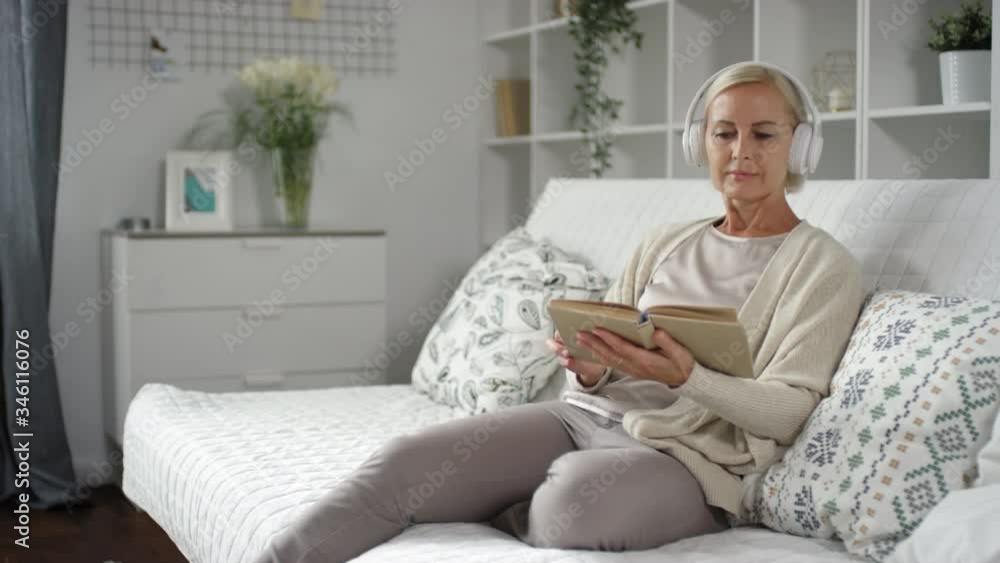 Tracking shot of blonde middle aged woman sitting on sofa at home, listening to music with wireless headphones and reading book Tracking shot of blonde middle aged woman sitting on sofa at home, listening to music with wireless headphones and reading book