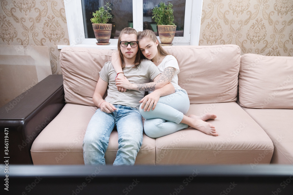 Attractive young long-haired couple hugging on a sofa at home in isolation watching tv. Concept of spending time with loved ones during a pandemic