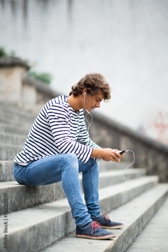 Full length side of young man sitting on steps listening to music with earphones and mobile phone Full length side of young man sitting on steps listening to music with earphones and mobile phone