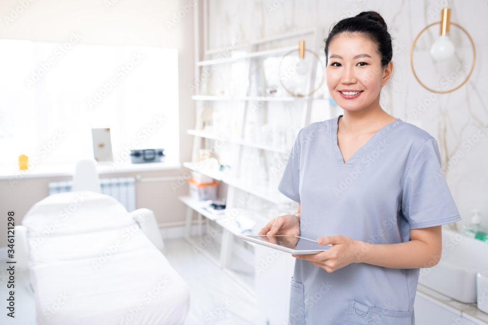 Portrait of positive attractive Asian cosmetologist in medical uniform using tablet in clean procedure room Portrait of positive attractive Asian cosmetologist in medical uniform using tablet in clean procedure room