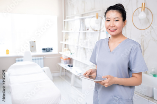 Portrait of positive attractive Asian cosmetologist in medical uniform using tablet in clean procedure room