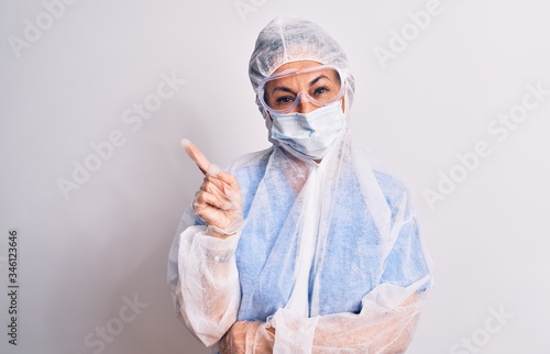 Middle age nurse woman wearing protection coronavirus equipment over white background with a big smile on face, pointing with hand and finger to the side looking at the camera.