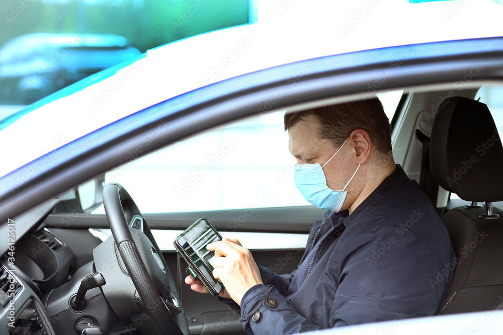A young man in a protective mask is sitting in a right-hand drive car and  calls on his mobile phone. Taxi driver. Coronavirus covid-19, pandemic. Virus protection concept