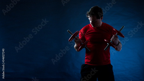 Young non-muscular man doing exercises with dumbbells on dark background