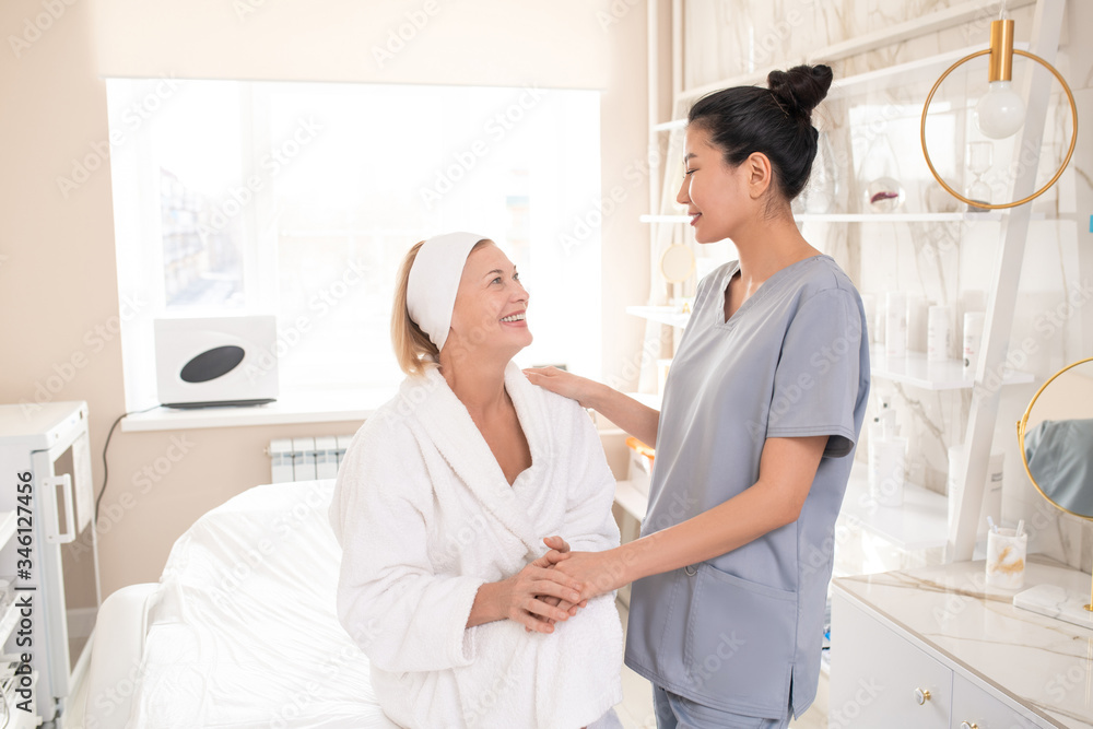 Satisfied mature beauty salon guest in robe sitting on procedure table and holding hand of Asian cosmetologist while thanking her after procedure Satisfied mature beauty salon guest in robe sitting on procedure table and holding hand of Asian cosmetologist while thanking her after procedure