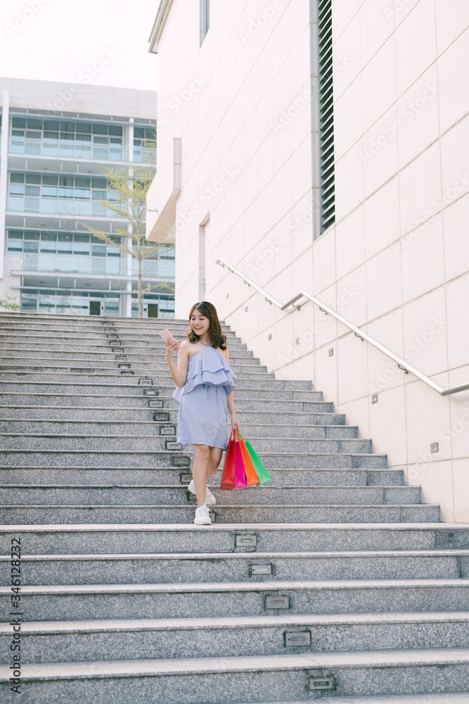 Happy young Asian woman using phone and going down stairs with colorful shop packages.