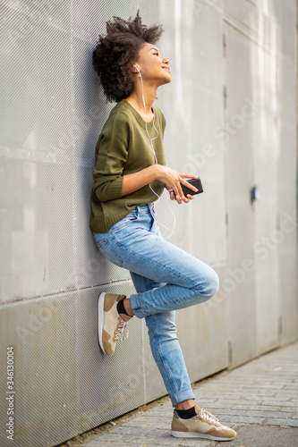 Bild auf Leinwand Full body smiling young african woman with mobile phone and earphones leaning ag