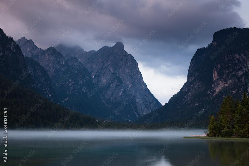 Lake Toblach with morning fog in the mountains of the Dolomite Alps during sunset, dramatic clouds in the sky, South Tyrol Italy.