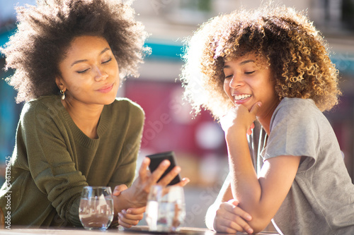 two happy young black women sitting at outdoor cafe looking at cellphone