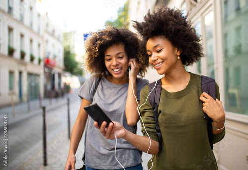 two female black friends walking in city with mobile phone listening to music with earphones photo