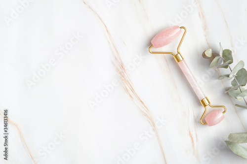 Rose quartz face roller and eucalyptus leaf on marble background, top view. Face massage tool, beauty treatment and facial skin care concept