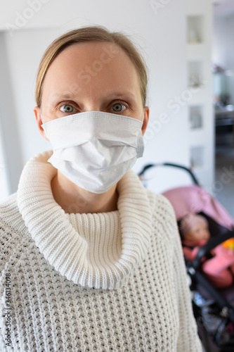 Serious woman wearing face mask, ready to walk with child, taking selfie at home, looking at camera with baby in pram in background. Corona virus pandemic concept