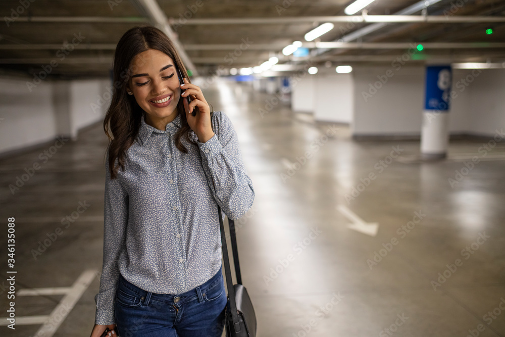 Fototapeta premium Portrait of young business woman with smart phone in the underground car parking of the new residential building. Businesswoman in underground garage. Elegant woman using smartphone in parking garage