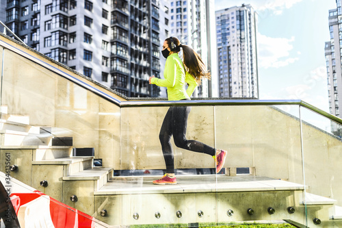 Healthy habits during quarantine. Full length young athlete woman with medical mask on the face in sports clothing is running up stairs while exercising outside