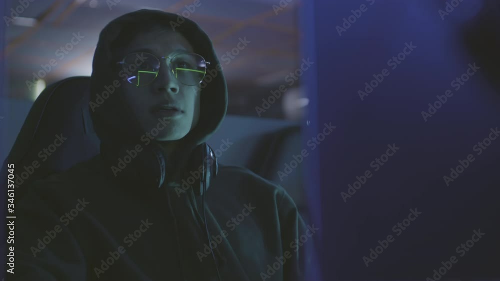 Lockdown portrait of young Asian man wearing eyeglasses and hoody sitting in dark computer club and playing Lockdown portrait of young Asian man wearing eyeglasses and hoody sitting in dark computer club and playing
