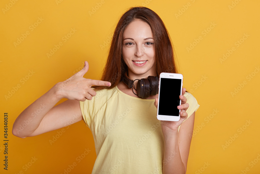 Closeup portrait of happy brunette female wearing casual t shirt, holding phone with blank screen and pointing at it with fore finger, having headphones around neck, isolated over yellow background. Closeup portrait of happy brunette female wearing casual t shirt, holding phone with blank screen and pointing at it with fore finger, having headphones around neck, isolated over yellow background.