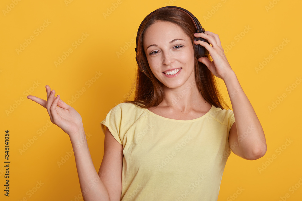 Fototapeta premium Indoor portrait of cheerful tender good looking model showing direction with forefinger, raising hands, looking directly at camera, having headphones, listening to music. People and chill concept.