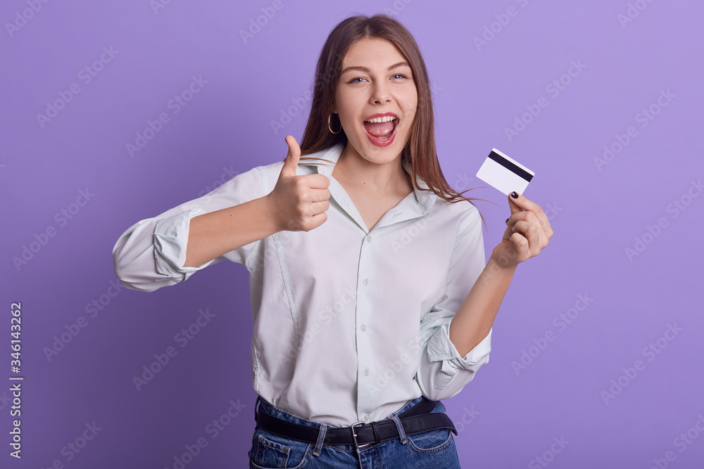 Picture of emotional sweet energetic young woman holding credit card, making gesture, thumb up, showing approval, opening mouth widely, wearing light shirt and jeans. People and money concept. Picture of emotional sweet energetic young woman holding credit card, making gesture, thumb up, showing approval, opening mouth widely, wearing light shirt and jeans. People and money concept.