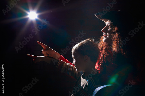 Ugle oman in red sweater and pirate hat with curly hair posing with small boy on black background. Actress mother and actor son in magical, fantastic, fairy-tale movie in dark studio and flash