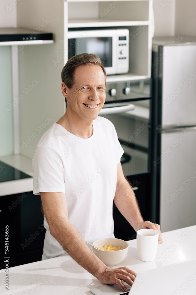 cheerful freelancer using laptop and touching cup near bowl with cornflakes cheerful freelancer using laptop and touching cup near bowl with cornflakes
