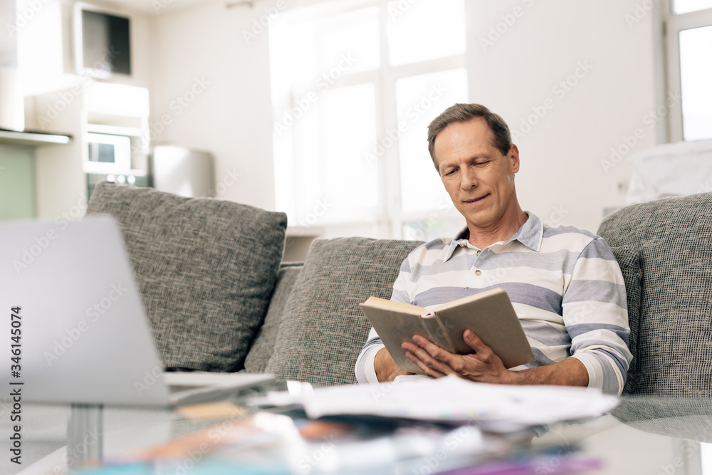 selective focus of handsome man smiling while reading book in living room selective focus of handsome man smiling while reading book in living room