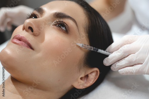 Close up of beautiful young woman is getting botox injection. Doctor is holding syringe near her eye area carefully.