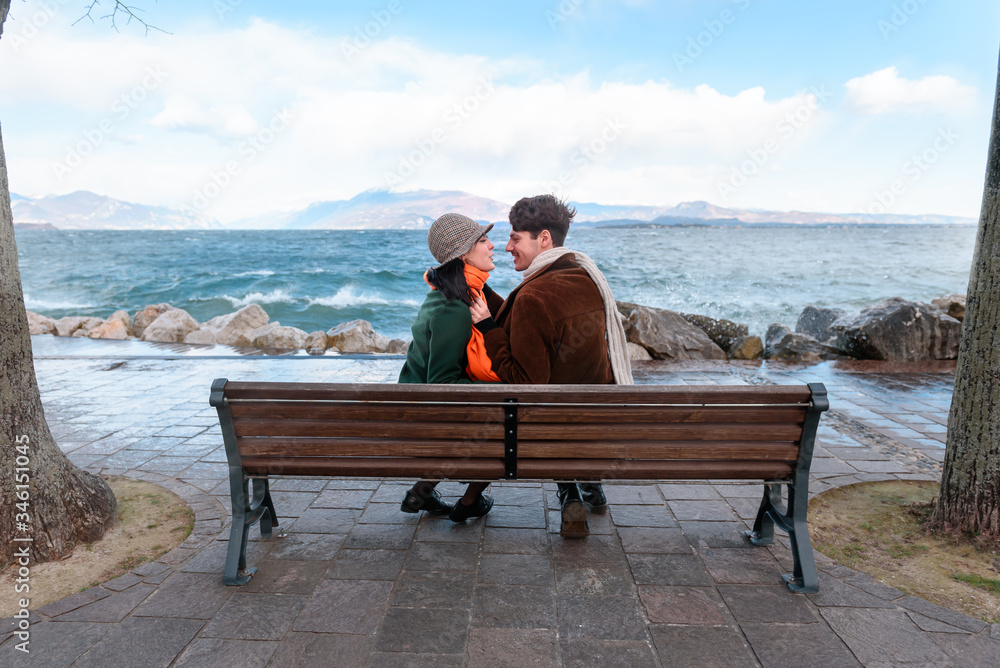 The young attractive girl and man are sitting on the bench. Young couple. Romantic mood. Couple in love. Street shot. Lake and mountains in the background