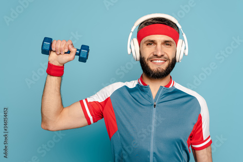 happy stylish sportsman in headphones exercising with dumbbell on blue background