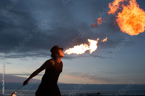 Obraz na plátně Young man with hat doing Fire breathing on the beach at dusk with sunset in the background