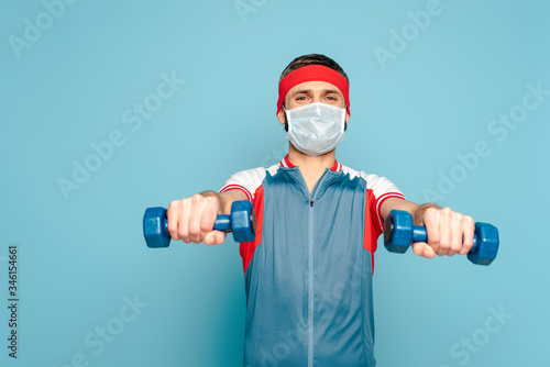 stylish sportsman in medical mask exercising with dumbbells on blue background