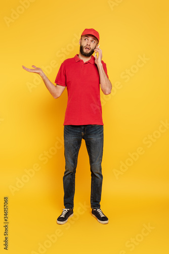 confused bearded delivery man in red uniform talking on smartphone on yellow