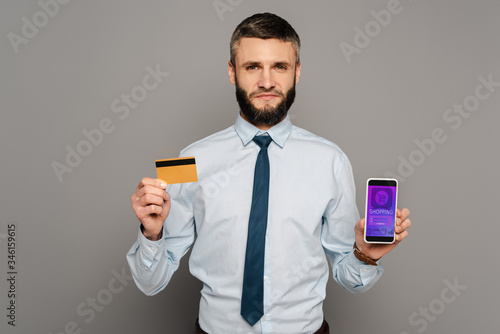 handsome bearded businessman holding credit card and smartphone with online shopping app on grey background