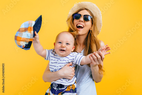 Happy beautiful mother in a hat and sunglasses, holds a little child, on an isolated yellow background. Travel, vacation, summer