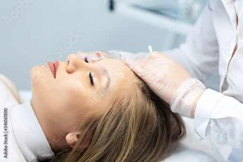 A young woman lies and master in a white uniform does eyebrow makeup in a beauty salon. The use of permanent makeup on brows. The wizard works with eyebrows. Semi-permanent makeup. Salon background.