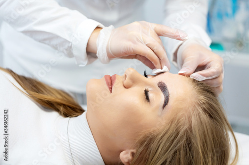 A young woman lies and master in a white uniform does eyebrow makeup in a beauty salon. The use of permanent makeup on brows. The wizard works with eyebrows. Semi-permanent makeup. Close up