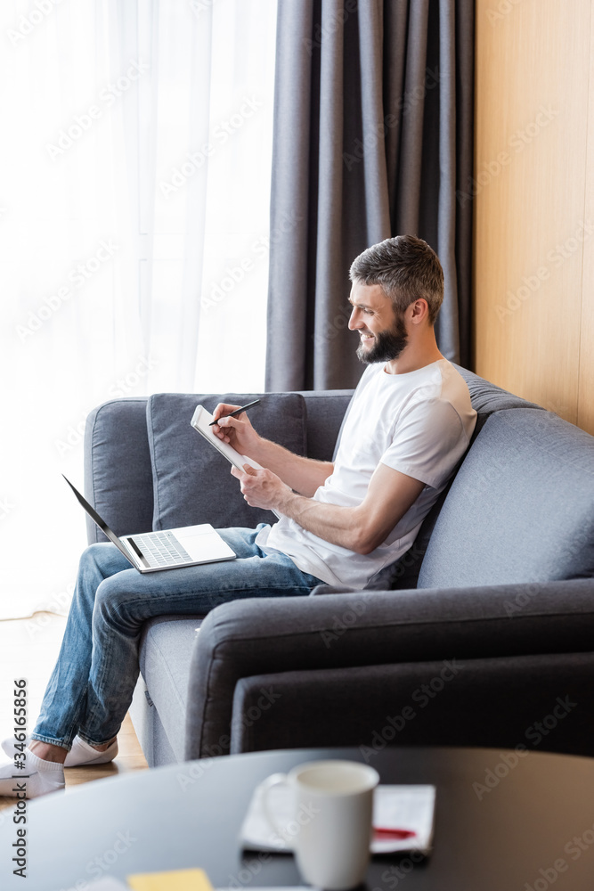 Selective focus of smiling teleworker working with laptop and notebook on sofa in living room Selective focus of smiling teleworker working with laptop and notebook on sofa in living room