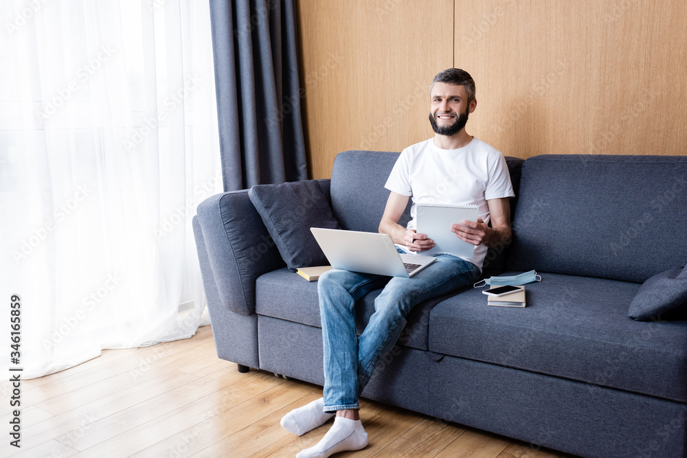 Smiling freelancer holding notebook near laptop, books and medical mask on couch at home