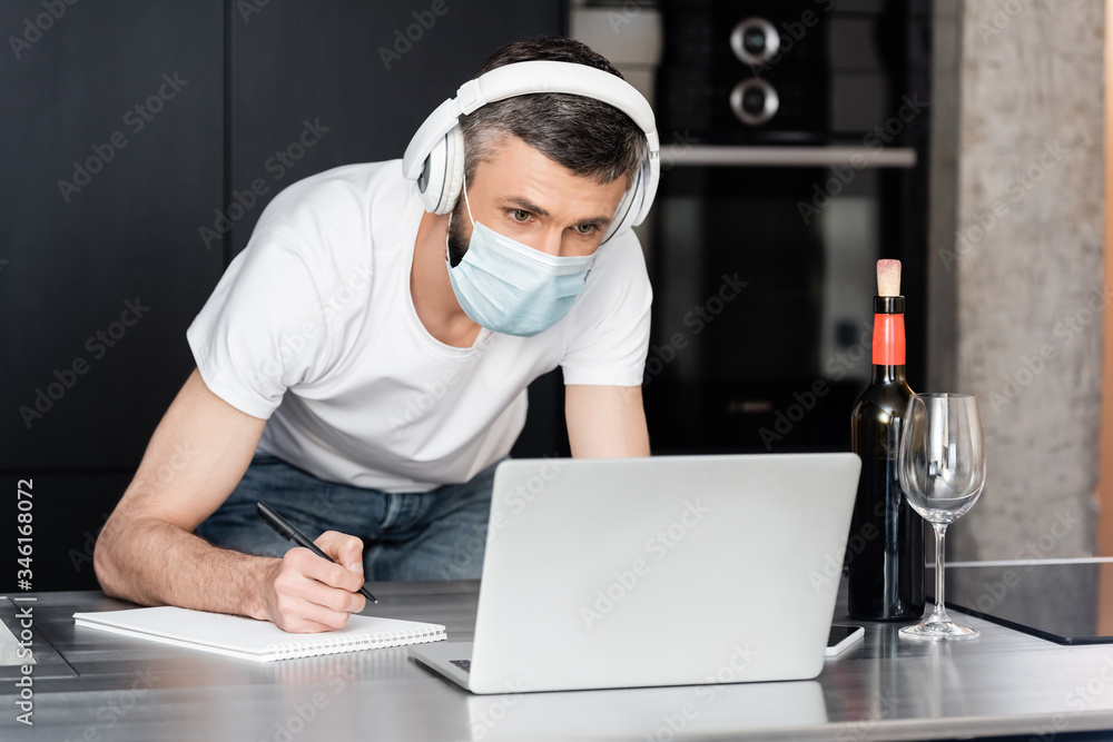 Teleworker in headphones and medical mask writing on notebook near laptop and wine on worktop in kitchen Teleworker in headphones and medical mask writing on notebook near laptop and wine on worktop in kitchen