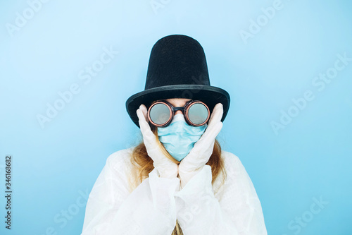 Creative girl in a white robe, medical mask, glasses and a hat on a blue background. Self-isolation during coronavirus quarantine. Epidemic covid-2019. © Stanislav