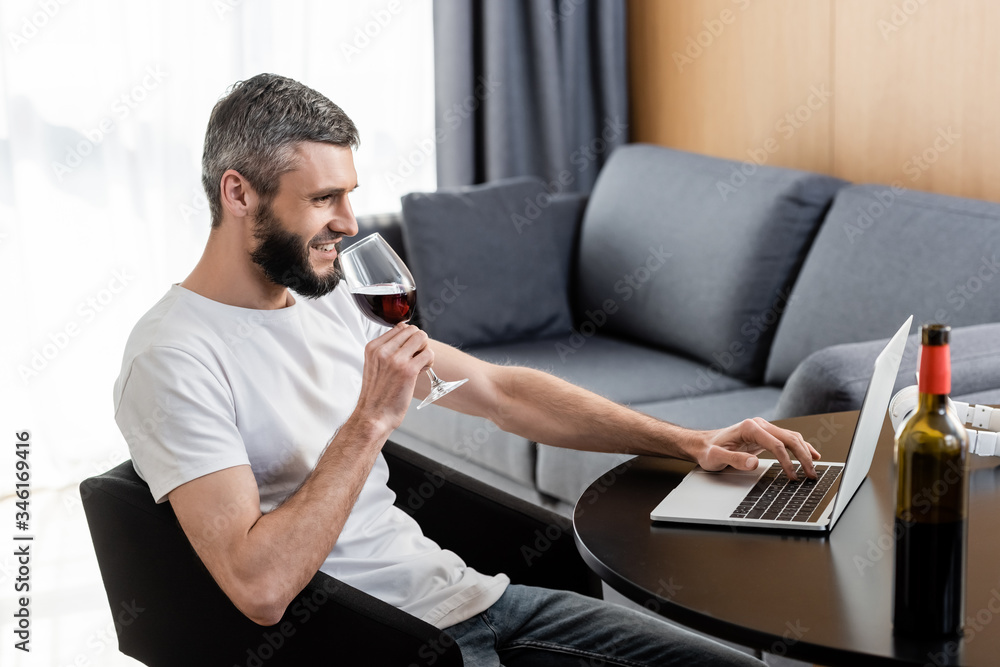 Side view of smiling freelancer drinking wine and using laptop in living room Side view of smiling freelancer drinking wine and using laptop in living room