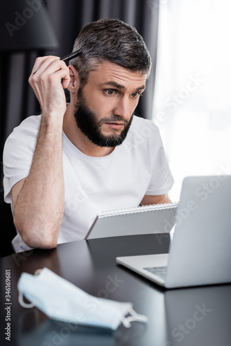 Selective focus of teleworker looking at laptop while holding notebook and pen near medical mask on table