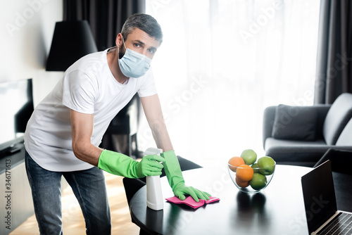 Man in medical mask and rubber gloves holding rag and detergent in living room