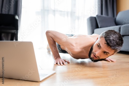 Selective focus of shirtless sportsman doing push ups near laptop on floor