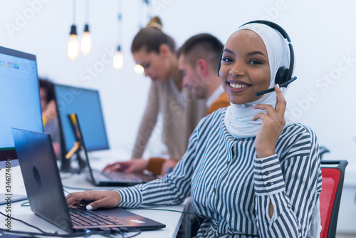Wall Mural African muslim female with hijab scarf customer representative business woman with phone headset helping and supporting online with customer in modern bright call centre