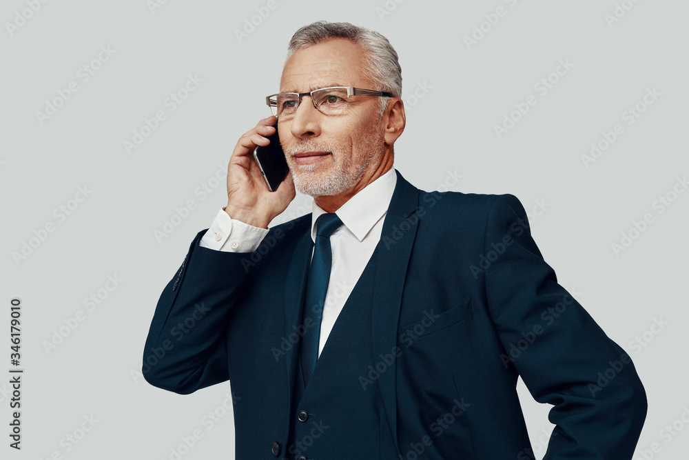 Handsome senior man in full suit talking on the phone while standing against grey background Handsome senior man in full suit talking on the phone while standing against grey background