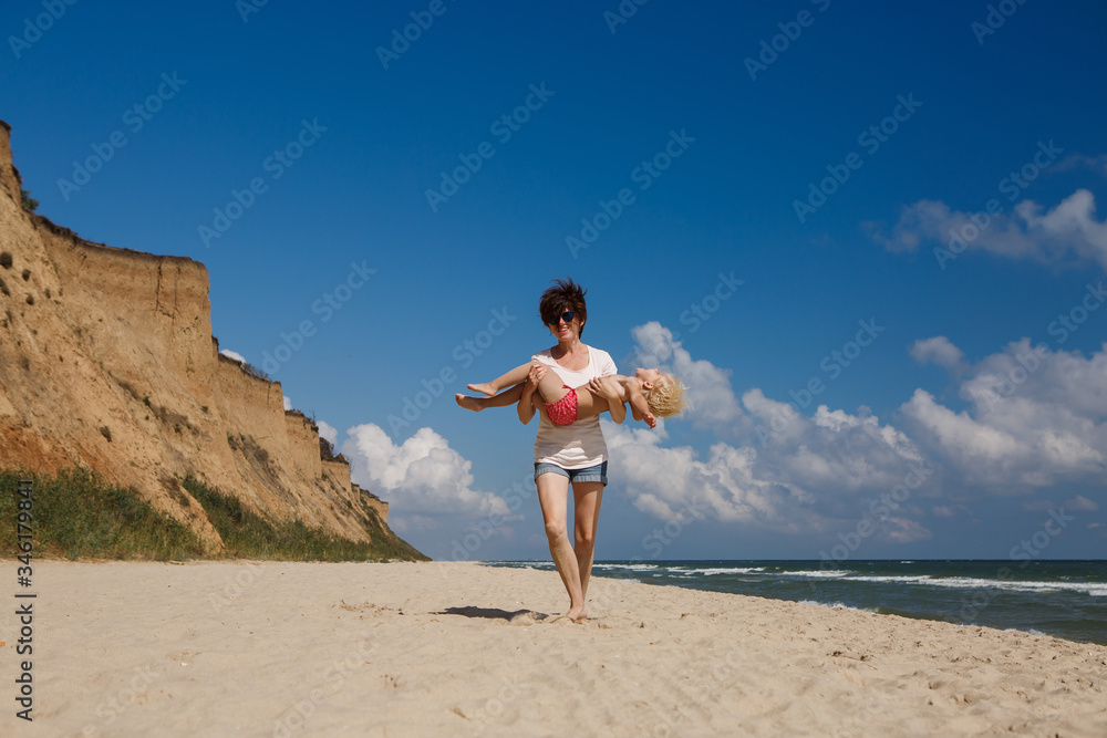 Little Cute Girl with Mother Plays with Mom on the Beach by the Sea in Summer. Portrait of a Perfect Happy Family.