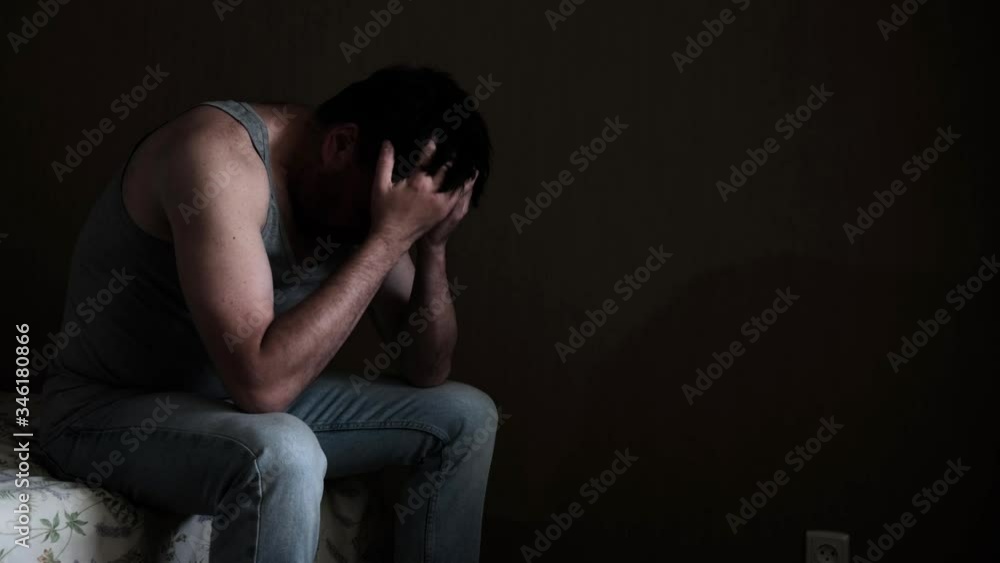Sad pensive depressed man sitting on a bed in a dark room Sad pensive depressed man sitting on a bed in a dark room