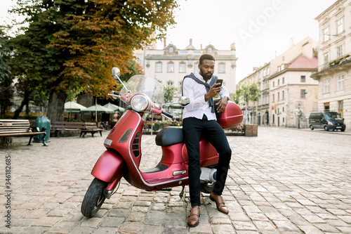 Full length portrait of fashionable African businessman wearing black pants and white shirt, leaning on red scooter in city center, writing message or reading news on his smartphone