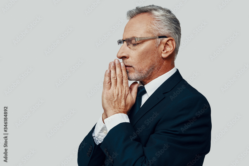 Handsome senior man in full suit keeping hands clasped while standing against grey background Handsome senior man in full suit keeping hands clasped while standing against grey background
